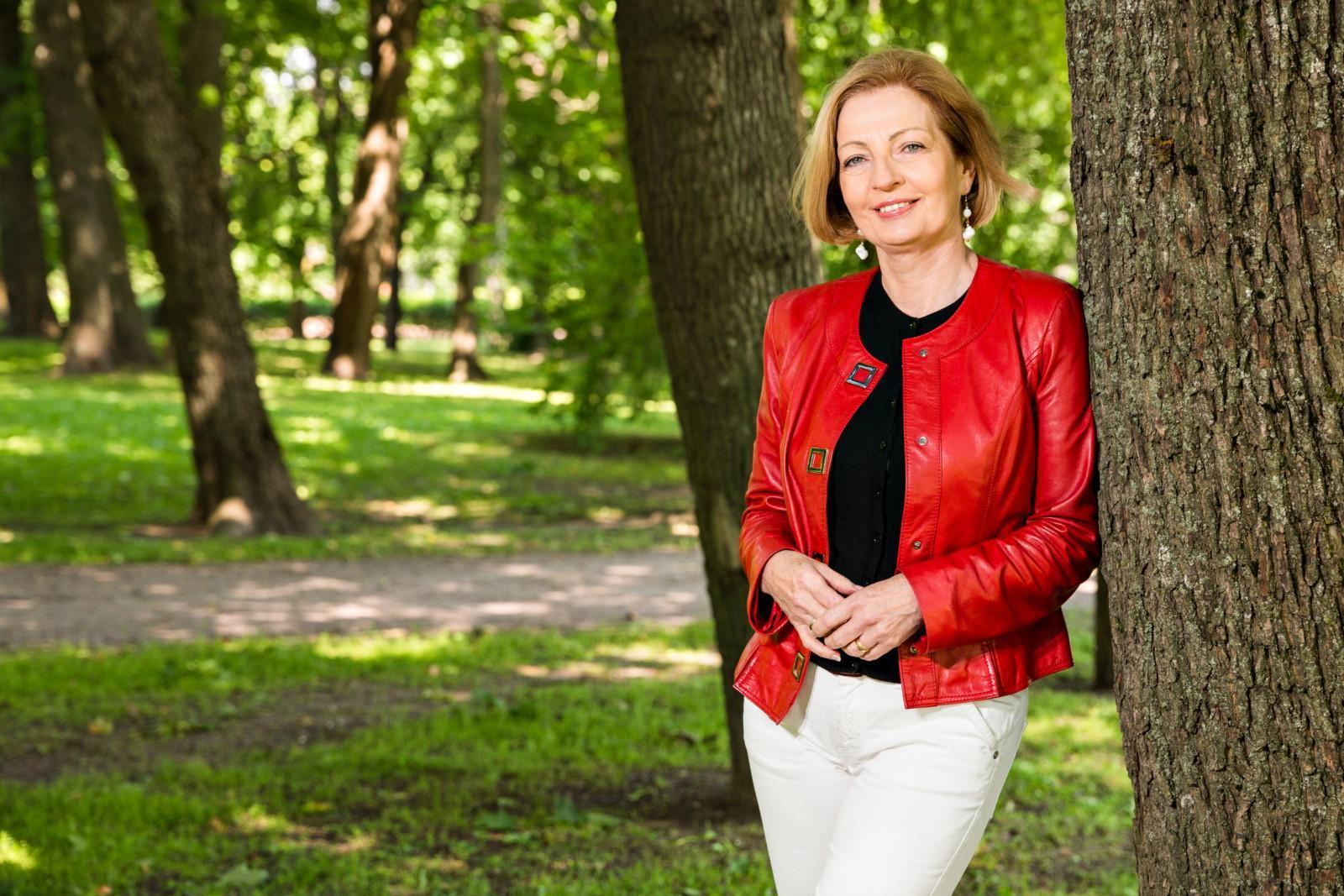 Hyvinvointilomat ry:n puheenjohtaja Anneli Kiljunen nojaa puuhun ja hymyilee aurinkoisessa puistossa.