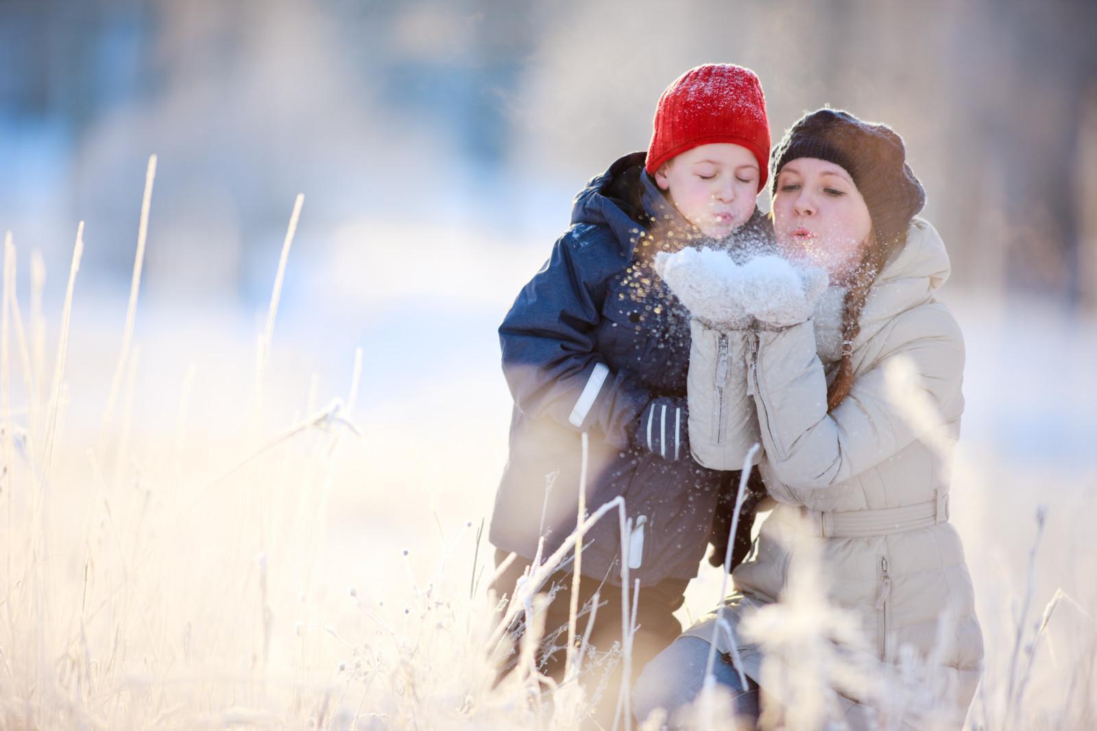 Nainen ja pieni poika puhaltavat lunta naisen käsistä lumisessa maisemassa.
