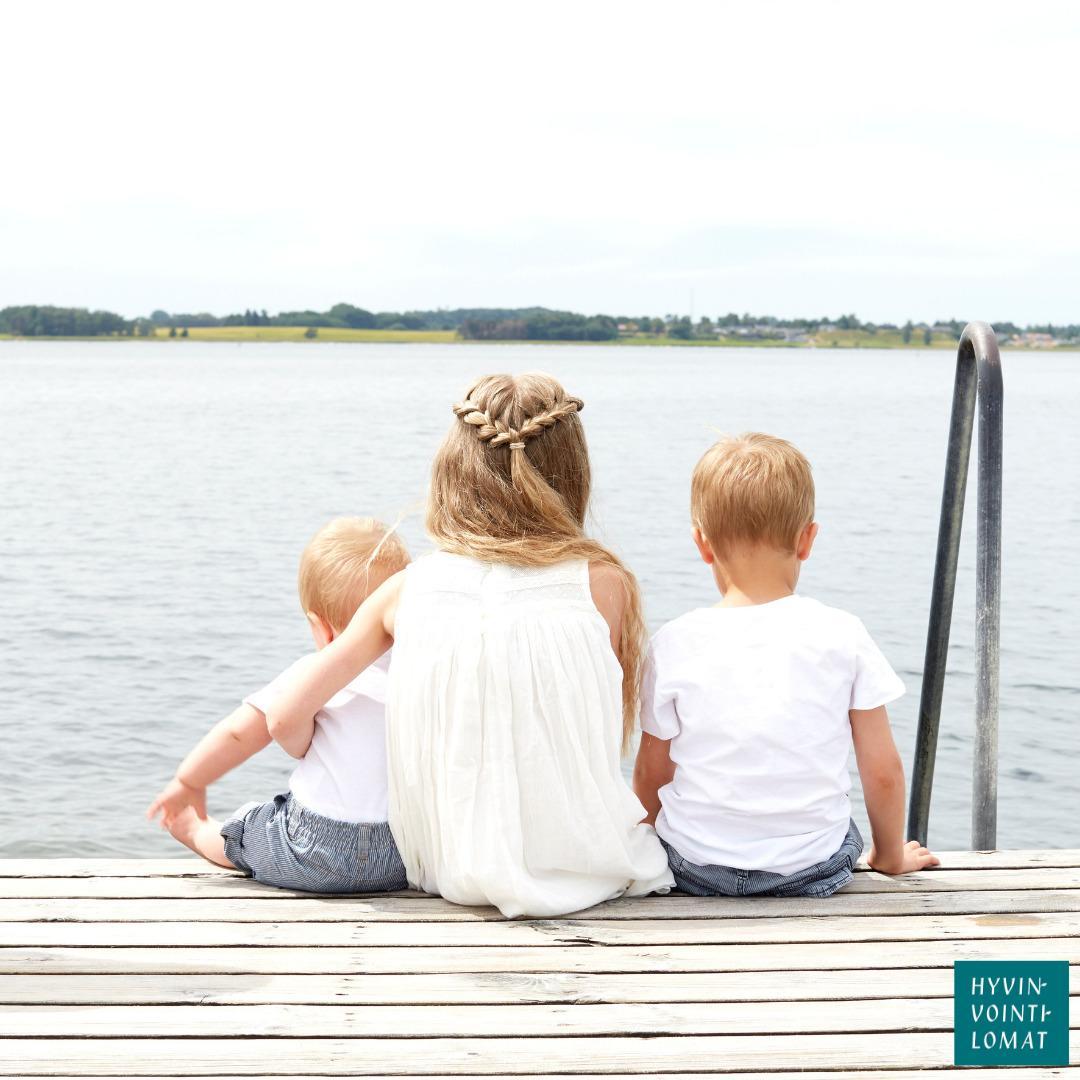 Valkomekkoinen tyttö istuu laiturilla järvi- tai jokimaisemassa. Tyttö pitää vasemmassa kainalossaan valkopaitaista ja harmaahousuista vauvaa. Tytön oikealla puolella istuu valkopaitainen ja farkkuhousuinen lapsi.