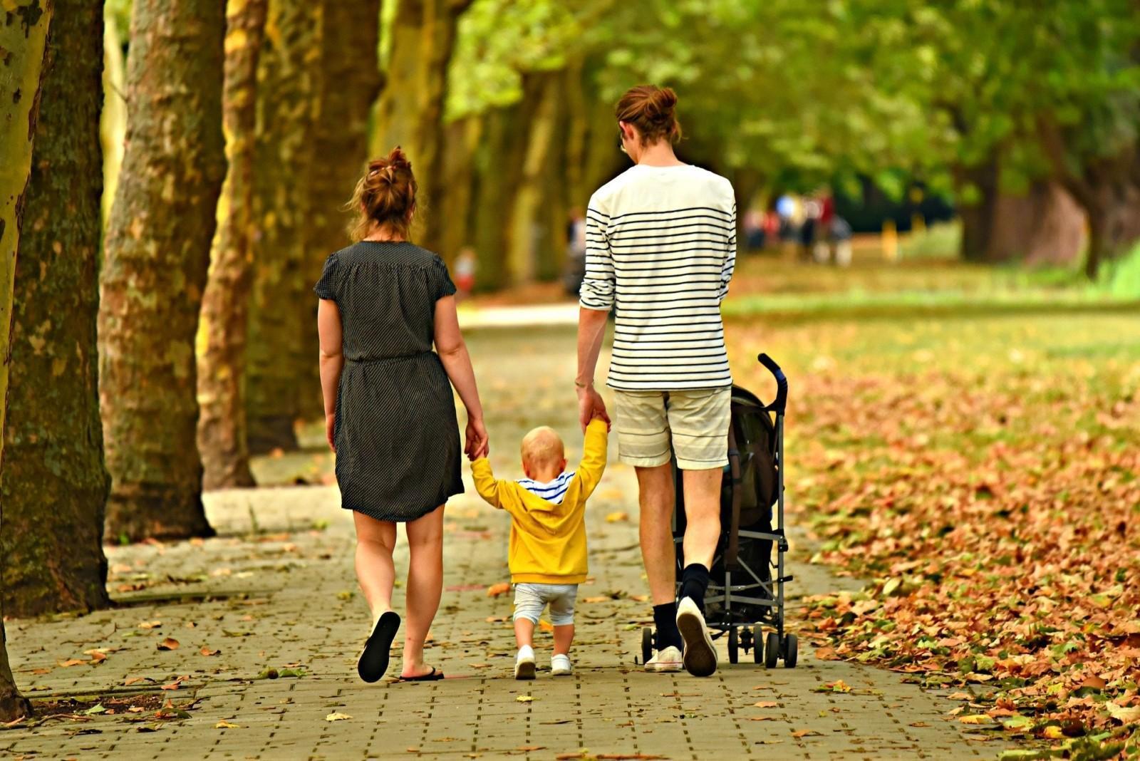 Nainen, mies ja pieni lapsi kävelevät käsi kädessä syksyisessä puistossa. Mies työntää lastenvaunuja.