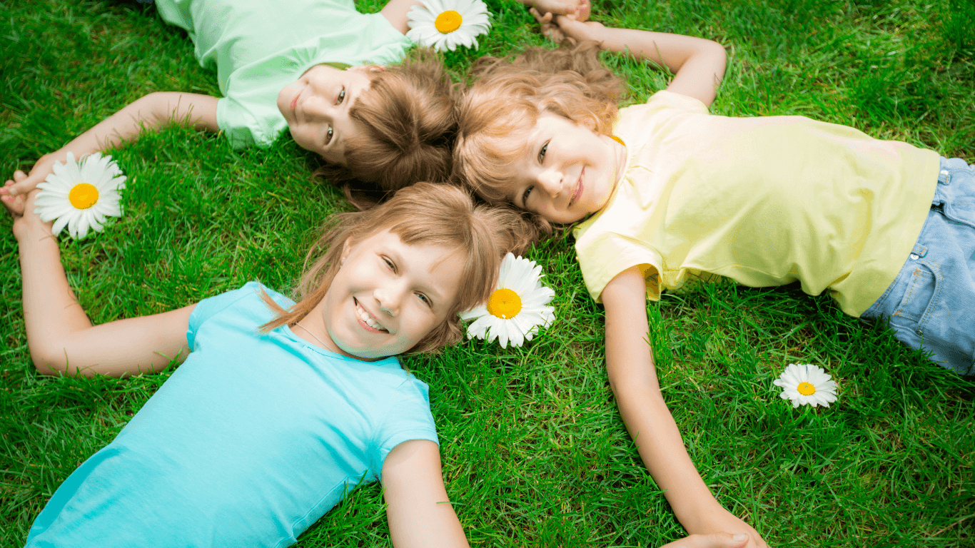 Kolme lasta makaa selällään nurmikolla toistensa käsistä kiinni pitäen. Lasten ympärillä on neljä päivänkakkaraa.