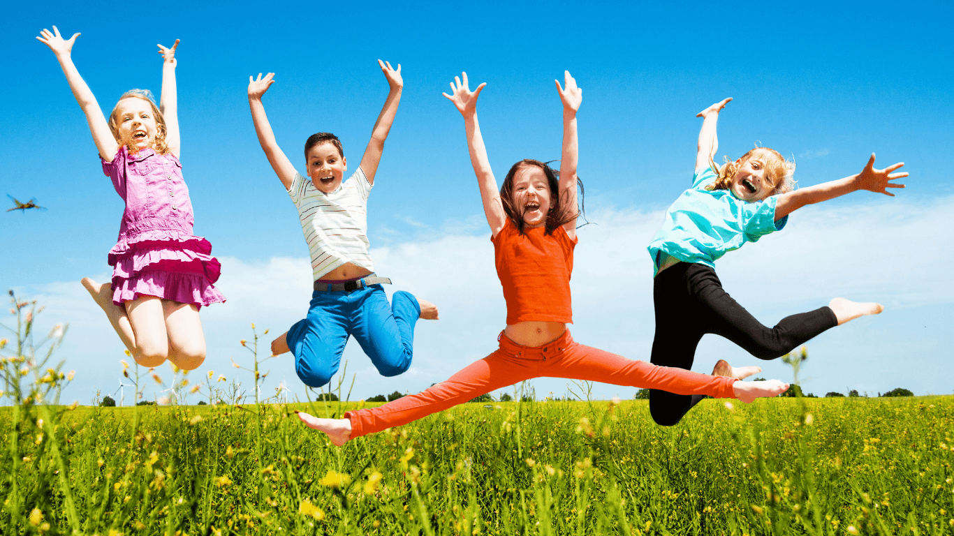 Neljä lasta hyppää iloisina ilmassa vihreällä niityllä kirkkaan sinisen taivaan alla, kädet ylhäällä ja värikkäät vaatteet päällä.