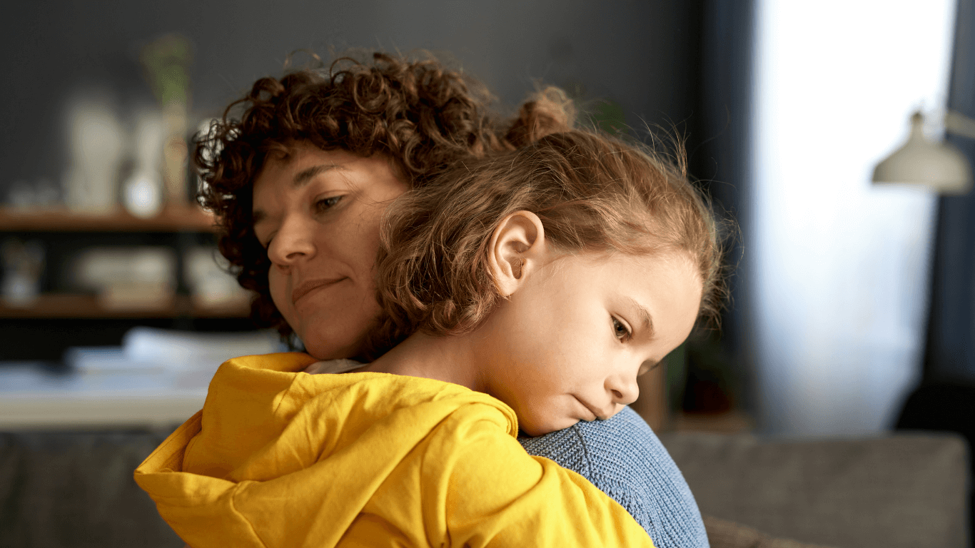 Äiti pitää sylissään alakuloisen oloista lasta kodin rauhallisessa ympäristössä. Lapsi nojaa äitiin ja katsoo mietteliäänä alaspäin.