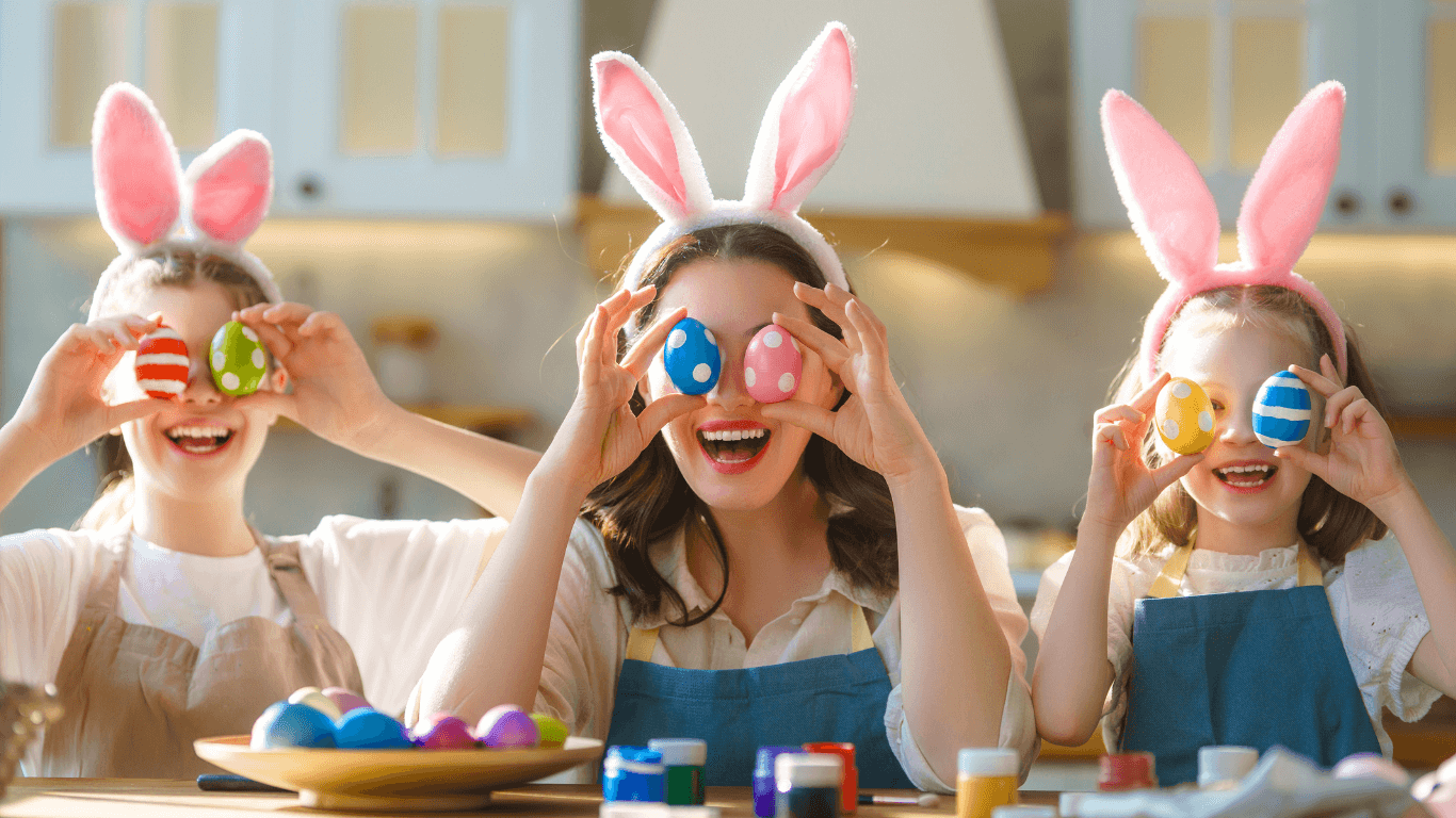 Kolme lasta pupunkorvat päässä istuu pöydän ääressä keittiössä ja pitelee värikkäitä pääsiäismunia silmiensä edessä kuin kiikareita. Pöydällä on maalipurkkeja ja lisää koristeltuja munia, ja lapset nauravat iloisesti.