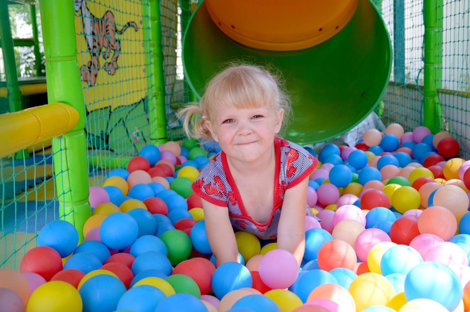 Vaaleahiuksinen pieni tyttö pallomeressä, jossa on sinisiä, punaisia ja keltaisia palloja. Taustalla näkyy vihreä liukumäki.