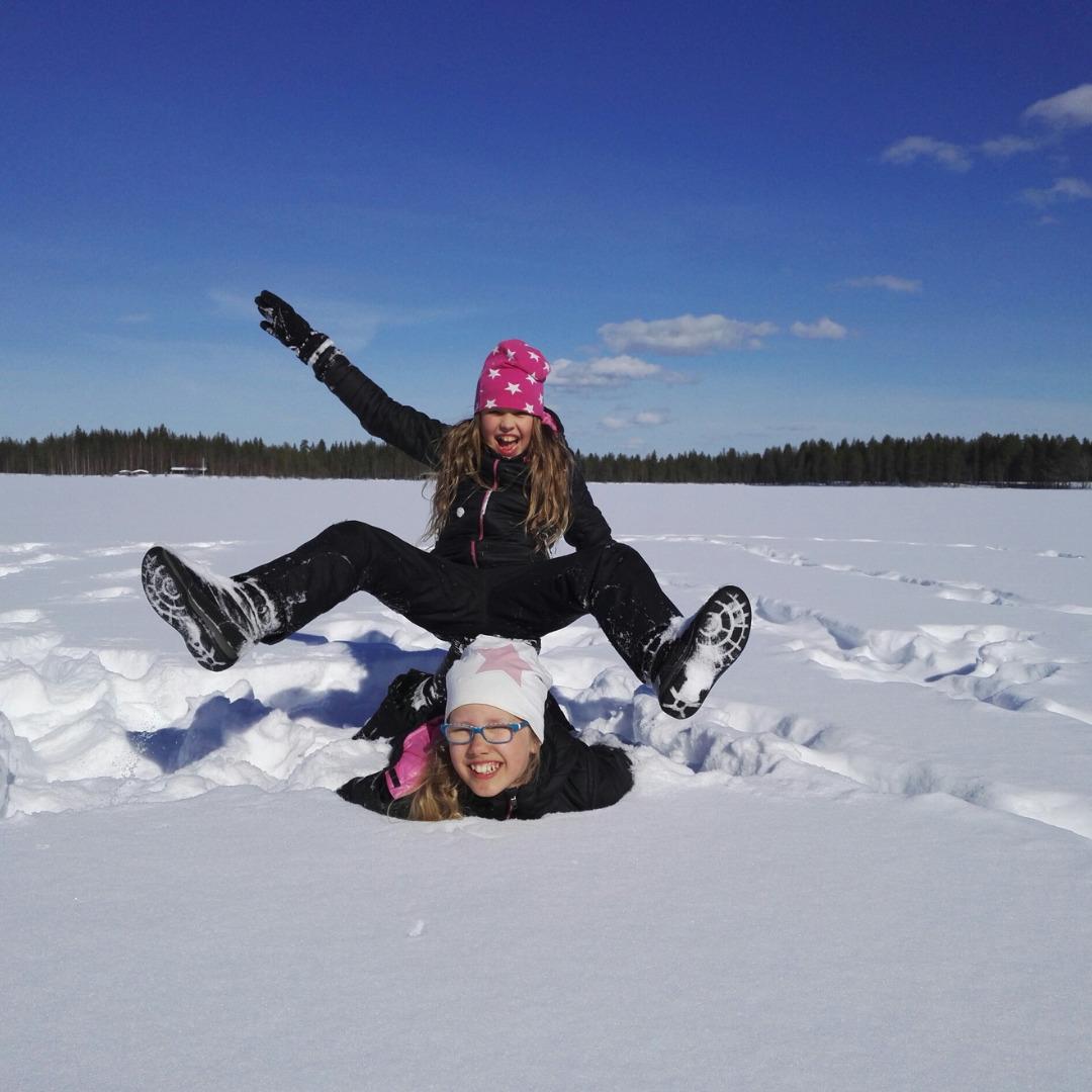 Kaksi tyttöä nauraa lumisessa maisemassa. Toinen makaa vatsallaan maassa ja toinen istuu hänen selässään jalat ja toinen käsi ylhäällä. Molemmat nauravat.