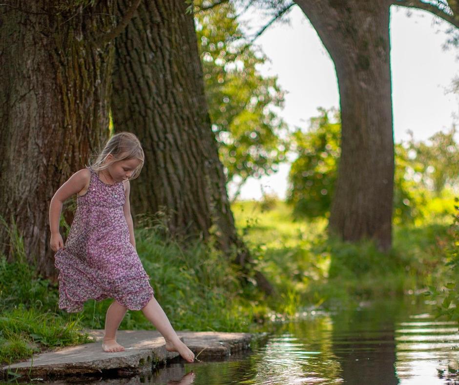 Pieni tyttö seisoo kannon päällä ja kastaa vasemman jalan varpaitaan veteen. Taustalla kesäinen metsä ja puita. Tytöllä on päällään vaaleanpunainen kesämekko.