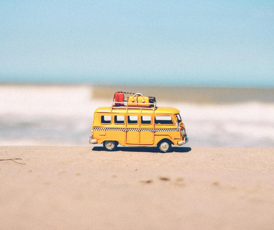 Keltainen Kleinbuss -mallinen keltainen pikkuauto hiekkarannalla. Auton katolla on pieniä erivärisiä matkalaukkuja.