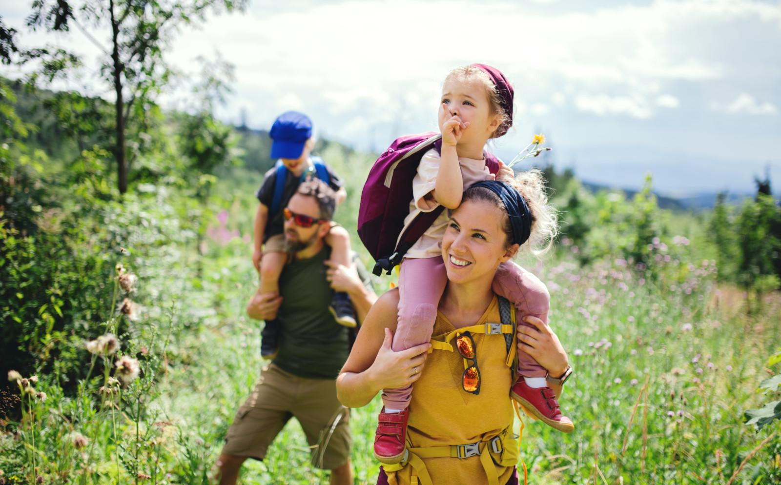 Mies kantaa olkapäillään pientä poikaa ja nainen pientä tyttöä. He ovat retkeilemässä kesäisessä luonnossa.